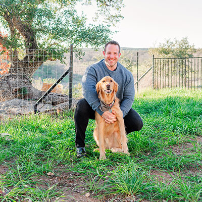 Smiling man squatting and hugging a golden retriever in a grassy field, with trees and a wire fence in the background on a sunny day.